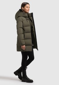 Zelená péřová bunda s kapucí, má zipový uzávěr, boční kapsy a prošívaný design. Nosená s černými legínami a kotníkovými botami.