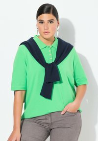 Zielona bawełniana koszulka polo z krótkimi rękawami i kołnierzykiem, z granatowym swetrem przewiązanym na ramionach. Szare dopasowane spodnie.