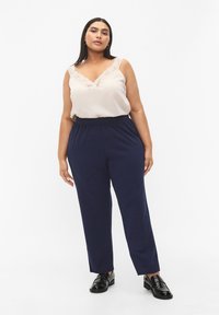 Light beige camisole with lace trim, paired with navy blue tapered pants. Model wears black, shiny lace-up shoes. Simple, modern design.