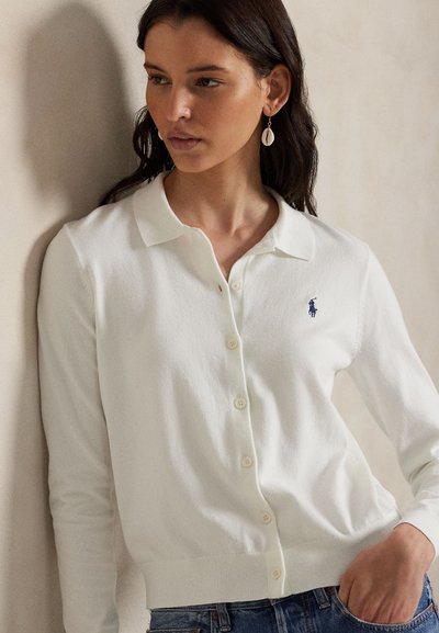 Krem jopica z mehko teksturo, z ovratnikom, zapiranjem na gumbe in majhno modro logotipom na levi strani prsnega koša.