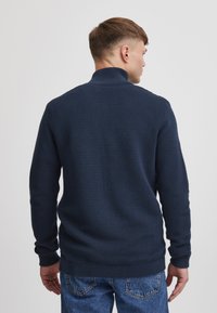 Granatowy prążkowany sweter z wysokim kołnierzem, wykonany z teksturowanego dziania, z długimi rękawami i dopasowanymi mankietami, zestawiony z niebieskimi dżinsami.