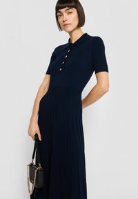 Tumši zilas adītas kleitas ar īsām piedurknēm, kurai ir apkakle un piecas zelta pogas pie kakla izgriezuma. Svārkiem ir vertikālas krokas, un tos papildina neliela melna soma.