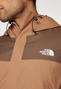 Veste à capuche marron avec un panneau sur la poitrine en marron plus foncé contrastant. Logo blanc affiché de manière visible sur le panneau, avec un cordon de serrage ajustable.