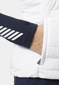 Mano en el bolsillo de una chaqueta blanca acolchada, con manga azul marino que tiene rayas diagonales blancas en el puño, vista en primer plano.