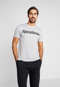 Grå t-skjorte med korte ermer som har en stor svart "Reebok"-logo over brystet, brukt med sorte bukser. Glatt stofftekstur.