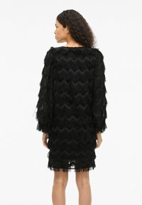 Robe frangée noire avec texture ondulée, manches longues et encolure arrondie. Le tissu est transparent, révélant un design à franges superposées.