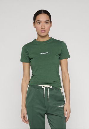 Ung kvinde med mørkt hår iført en grøn Pegador t-shirt og matchende joggingbukser med snor, stående foran en ensartet lys baggrund.