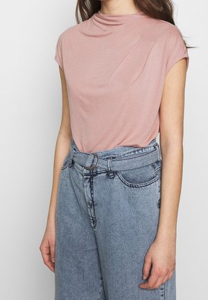 Frau trägt ein hellrosa kurzärmeliges Oberteil, das in hoch taillierte hellblaue Jeans mit passendem Jeansgürtel gesteckt ist.