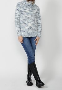 Pull en maille bleu clair avec un col cheminée, présentant un motif texturé. Associé à un jean bleu et des bottes noires à genoux avec des semelles épaisses.