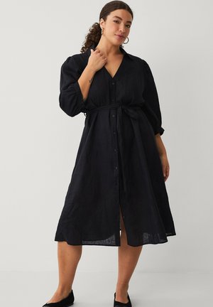 Kvinde med krøllet hår iført en sort midikjole med knapper, puffede ærmer og bånd i taljen, stående mod en ensfarvet lys baggrund.