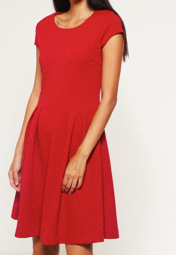 Femme portant une robe rouge à manches courtes, longueur genou, avec un corsage ajusté et une jupe plissée sur un fond uni.