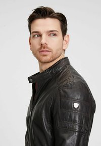 Cazadora de cuero negro con cuello alzado, detalles acolchados en los hombros y una placa con logo en la manga izquierda. Textura suave y diseño ajustado.