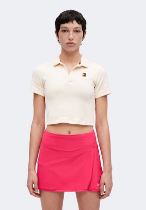 Jeune personne aux cheveux courts et foncés porte un polo court couleur crème et une jupe de tennis sportive rose vif, debout devant un fond uni.