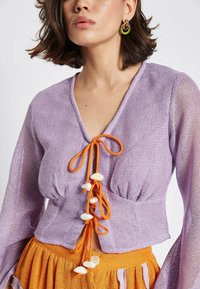 Lavender mesh top with orange ties and shell accents, featuring long sleeves and a v-neckline. Paired with an orange skirt.