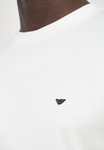 Λευκό βαμβακερό κοντομάνικο μπλουζάκι με μικρό μαύρο κεντητό λογότυπο στο αριστερό στήθος. Λείο υφασμάτινο με στρογγυλή λαιμόκοψη.