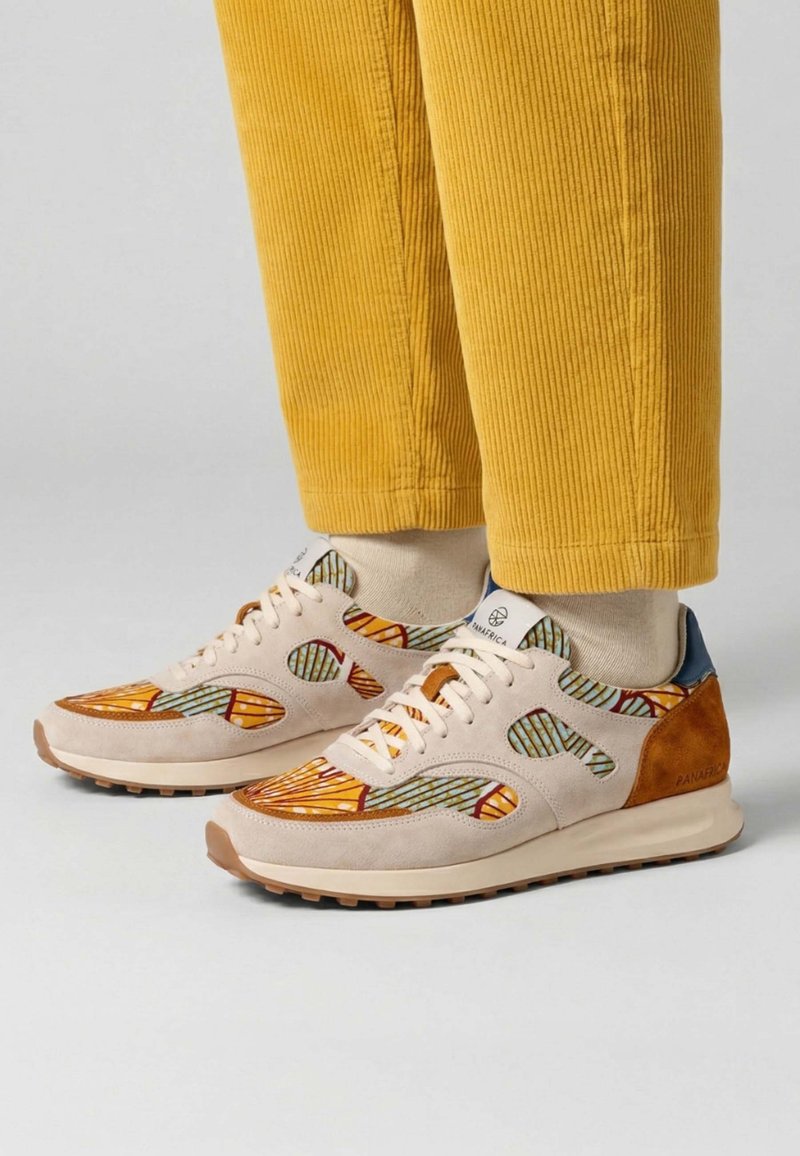 Personne portant un pantalon en velours côtelé jaune et des baskets beiges avec des empiècements en tissu coloré à motifs, debout sur un fond clair uni.