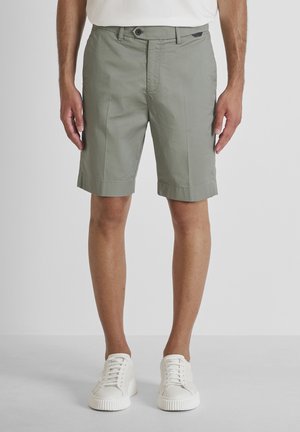 Hombre con pantalones cortos gris claro hasta la rodilla y zapatillas blancas, de pie contra un fondo liso y claro.