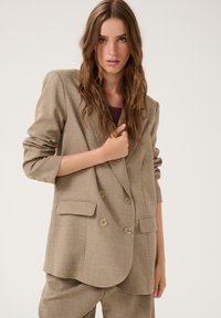 Blazer beige croisé avec un col à revers, en tissu texturé, quatre boutons et deux poches avant. Manches trois-quarts.