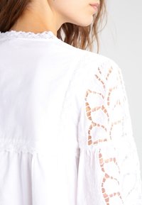 Blouse blanche avec des manches transparentes brodées, un délicat ourlet en dentelle le long de l'encolure, et une coupe ample.