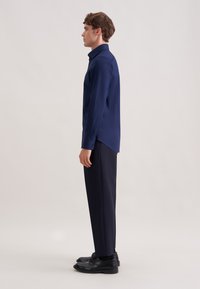 Camicia blu navy a maniche lunghe con colletto, dettagli sulle cuciture laterali, indossata con pantaloni scuri e scarpe nere, ripresa di profilo su uno sfondo chiaro.