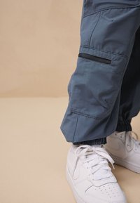 Pantaloni cargo blu con polsini elasticizzati e tasche laterali, abbinati a scarpe da ginnastica bianche con punta traforata e lacci.