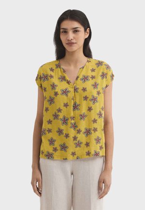 Kobieta o długich ciemnych włosach, ubrana w żółtą bluzkę z krótkim rękawem w zielono-różowy kwiatowy wzór oraz jasnobeżowe spodnie, stoi na jasnym tle.