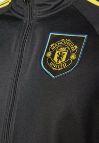 Melna jaka ar rāvējslēdzēju, uz tās kontrastējošs dzeltena un zila Mančestras United logo. Izgatavota no teksturēta materiāla ar rāvējslēdzēja elementiem.