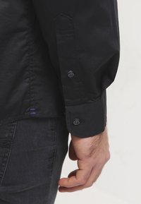Close-up van de pols en hand van een persoon die een zwart gewatteerd shirt met knopen en donkere spijkerbroek draagt, tegen een effen lichte achtergrond.