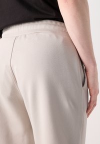 Pantalons taper aux teintes grises claires avec une texture lisse, ceinture élastique, poches latérales et détails de couture. Gros plan arrière.