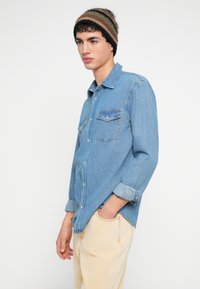 Jasnoniebieska koszula dżinsowa z zapięciem na guziki, dwoma kieszeniami na piersi i podwiniętymi rękawami, zestawiona z kremowymi spodniami. Wełniana czapka w paski.