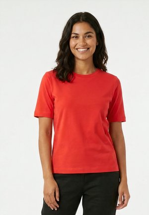 Femme aux cheveux foncés mi-longs portant un t-shirt rouge à manches courtes et un pantalon noir, souriante sur fond blanc.