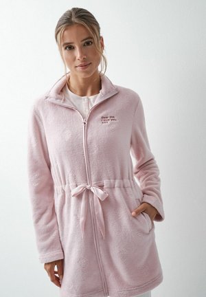 Jeune femme portant une veste polaire zippée rose pâle avec une ceinture à la taille et un texte brodé sur la poitrine, debout devant un fond blanc.