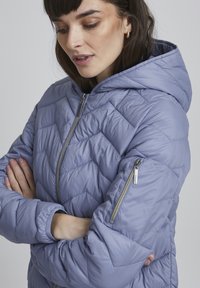 Giacca a puffo leggera con cappuccio in morbido lavanda, con trapuntatura a chevron, zip frontale e tasche laterali. Texture liscia, maniche elastiche.
