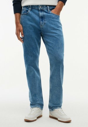 Personne portant un jean droit bleu clair et des baskets blanches, debout avec une main dans la poche sur un fond uni.