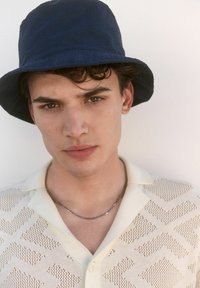 Cappello a secchiello blu navy abbinato a un polo lavorato a maglia di un beige chiaro con un motivo a rete geometrica. Maniche corte e una collana in argento.