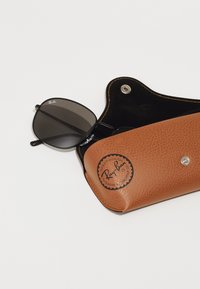 Ochelari de soare Ray-Ban cu lentile negre într-un toc din piele texturată de culoare maro, cu un detaliu rotund cu logo și o închidere tip clapetă. Interiorul este de culoare neagră.