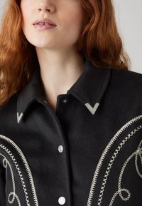 Melnas vilnas krekla jaka ar sudraba V formas apkakles akcentiem un sarežģītu balto izšuvumu gar priekšpusi vijīgās rakstās.