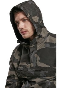 Zöld és szürke árnyalatokban kapható álcázó kapucnis dzseki, elülső zsebbel, állítható húzózsinórral és matt felületű textúrával.