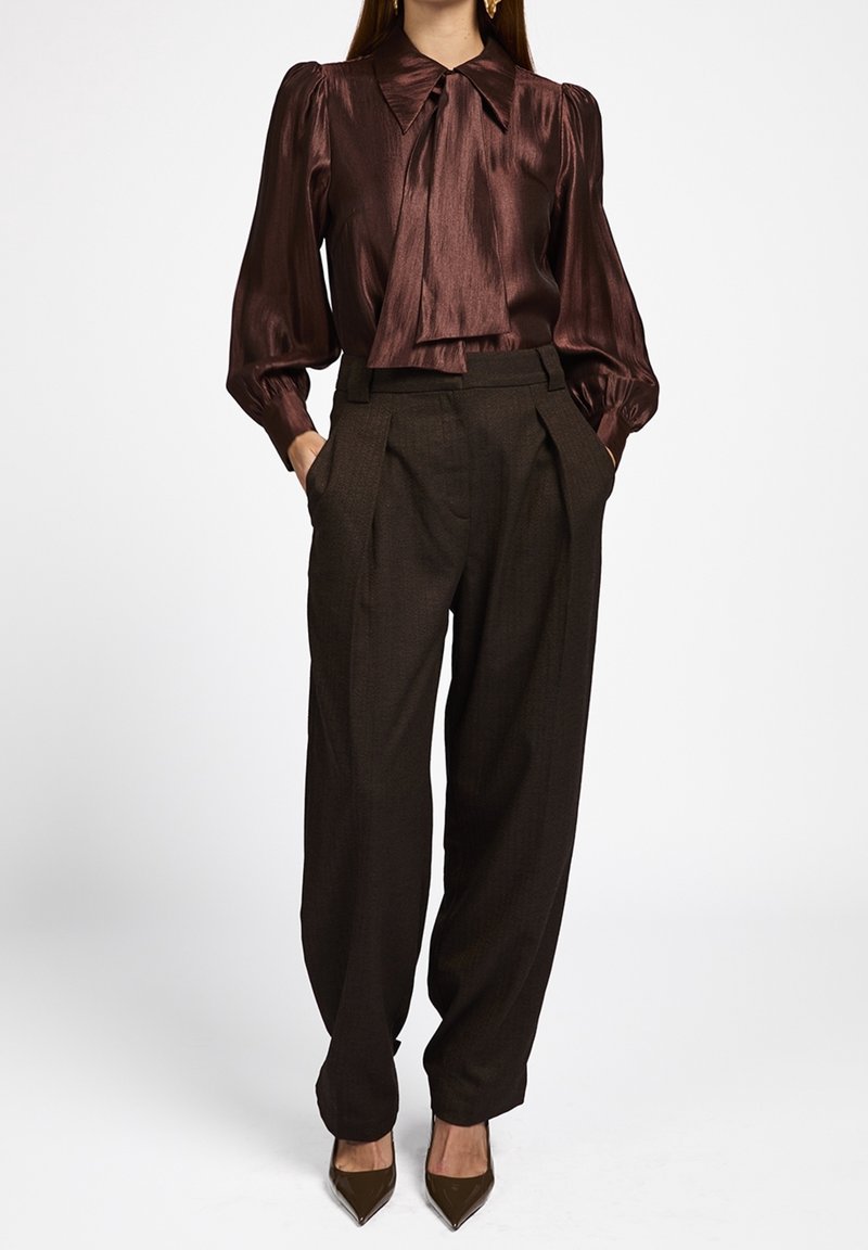 Femme portant un chemisier marron brillant à manches longues avec un grand col et un nœud, associé à un pantalon plissé marron foncé taille haute et des talons.
