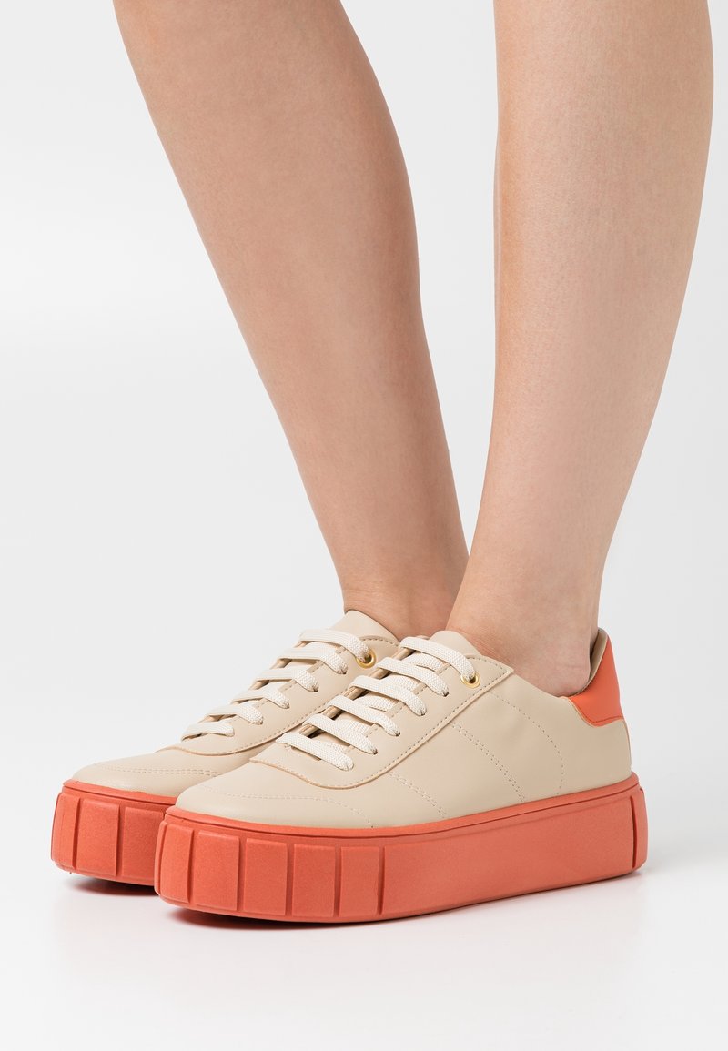 Baskets en cuir beige avec une semelle plateforme texturée orange. Comprend des lacets blancs et des œillets ronds dorés.
