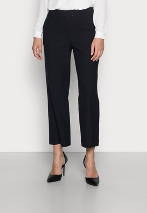 Kvinde iført sorte, skræddersyede, korte bukser med dobbelt knaplukning og sorte højhælede sko med spids tå, stående mod en ensfarvet baggrund.