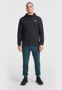 Haut noir avec une poche avant et un cordon de serrage réglable, associé à un pantalon turquoise avec un logo sur le côté, et des baskets noires.
