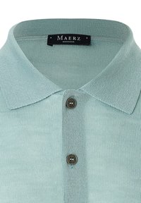 Ljusgrön stickad poloskjorta med klassisk krage och en mörk knapp vid halsen, med en strukturerad tygfinish.