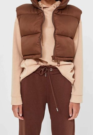 Personne portant un sweat à capuche beige avec cordons, un gilet sans manches matelassé marron court et non zippé, et un pantalon de jogging marron à cordons.