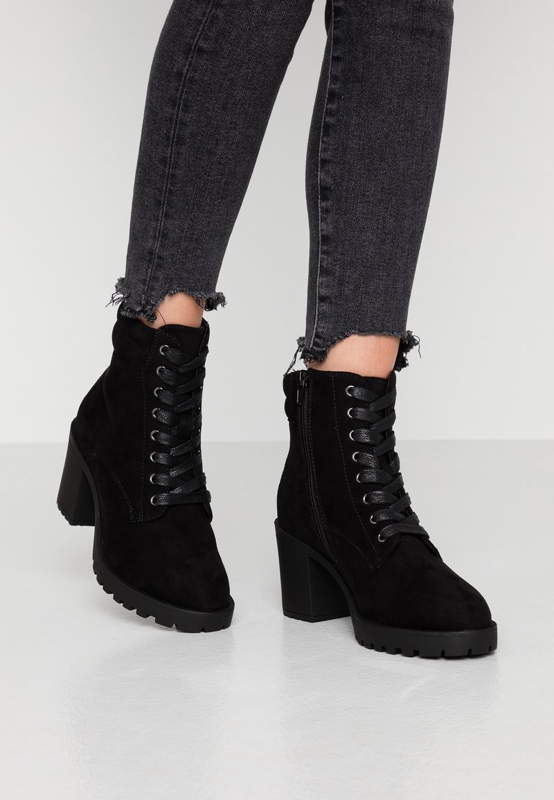 Bottines à lacets en daim noir avec talons épais portées avec un jean skinny gris foncé à ourlet effiloché sur fond blanc.