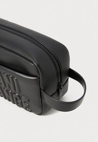 Črna usnjena torba z teksturirano površino, zadrgo in embosiranim detajlom "ANTI". Vključuje okroglo ročico za enostavno nošenje.