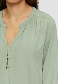 Blouse vert clair à manches longues avec un col rond, présentant une patte de boutonnage et des détails froncés aux épaules. Texture de tissu douce.