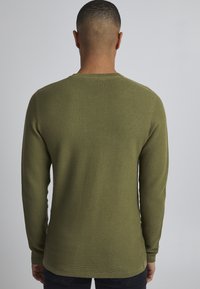 Pulover lung cu mânecă lungă de culoare verde măslin, cu un model de tricotaj texturat, decolteu rotund și o ușoară coadă la tiv. Vederea din spate evidențiază detaliile de cusătură.