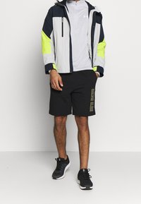 Herrenwindbreakerjacke in Grau, Marineblau und Neon-Gelb, kombiniert mit schwarzen Shorts, die den gelben "Calvin Klein"-Schriftzug tragen, und schwarzen Sportschuhen.