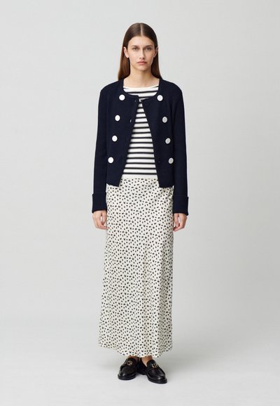 Cardigan côtelé marine avec de grands boutons argentés, top rayé et jupe blanche à pois noirs. Des mocassins noirs complètent la tenue.